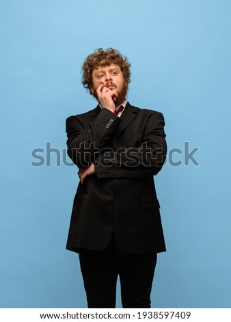 Thinking. Portrait of young hairy red-headed man in black business suit isolated over blue background. Concept of human emotions, facial expression, sales, business modern life. Copyspace for ad.