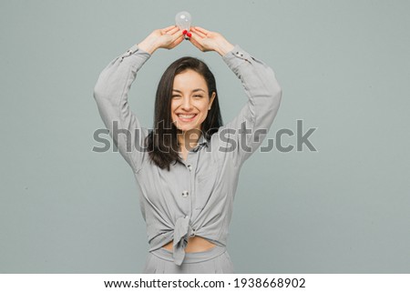 Photo of cute smiling female holds lamp over her head. Wears grey t-shirt, isolated grey color background