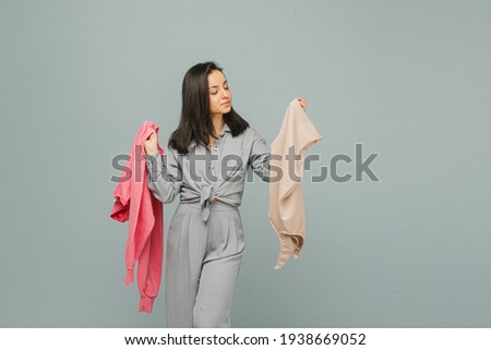 Photo of cute smiling female holds sweaters and look at one. Wears grey t-shirt, isolated grey color background
