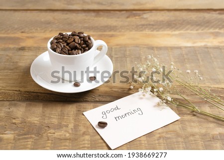 Coffee beans in a white cup with a card that reads "Good morning!" on a wooden background. Morning coffee concept. 