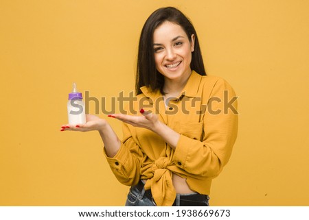 Photo of cute female holds baby bootle of milk. Wears yellow shirt, isolated yellow color background