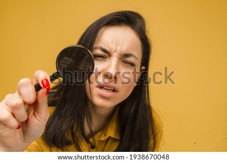 Photo of cute female holds magnifying glass and looking for something. Wears yellow shirt, isolated yellow color background