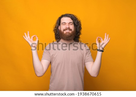 Portrait of young bearded hipster man making zen gesture over yellow background.