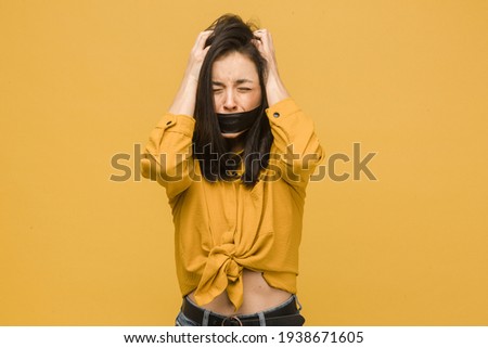 Concept photo of female victim with her mouth taped up, holds her head. Wears yellow shirt, isolated yellow color background