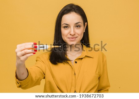 Concept photo of cute female in face mask holds thermometer. Wears yellow shirt, isolated yellow color background