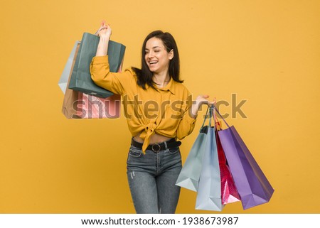 Concept photo of young female holds shopping bags, buying clothes. Wears yellow shirt, isolated yellow color background