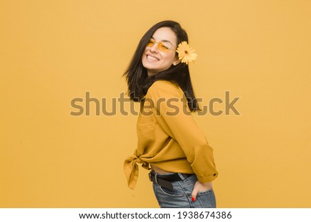 Concept photo of young female in glasses and flower in hair, feeling spring time. Wears yellow shirt, isolated yellow color background
