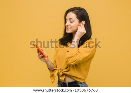 Close up photo of young female is using EarPods and smartphone. Wears yellow shirt, isolated yellow color background