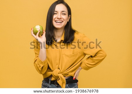 Concept photo of young female holds apple and smile, healthy food. Wears yellow shirt, isolated yellow color background