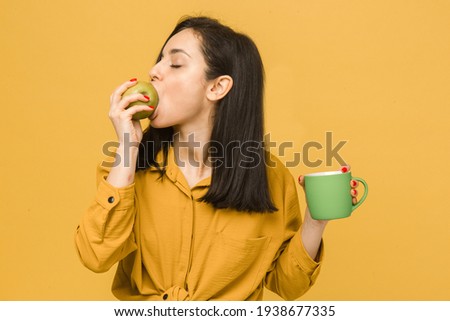 Concept photo of young female eating apple and drinking something. Wears yellow shirt, isolated yellow color background