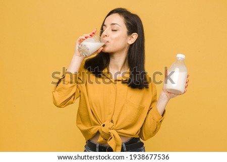 Concept photo of young female is drinking milk in glass. Wears yellow shirt, isolated yellow color background