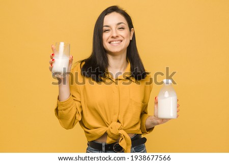 Concept photo of young female is holding glass and bootle of milk. Wears yellow shirt, isolated yellow color background