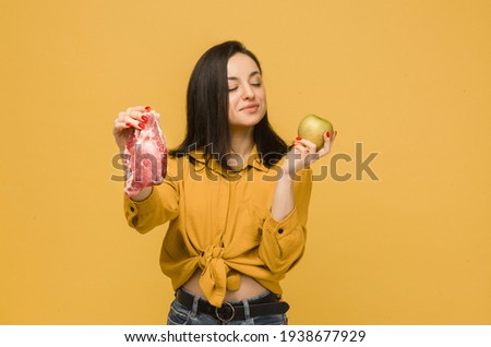 Concept photo of sweet female chooses between meat and apple. Wears yellow shirt, isolated yellow color background
