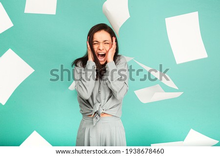 Photo of cute female in glasses is screaming, paper is flying around her. Wears grey shirt, isolated turquoise color background