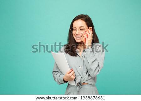 Photo of cute business lady in glasses holds papers and talking on phone. Wears grey shirt, isolated turquoise color background