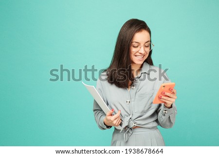 Photo of cute business lady in glasses holds papers and texting on phone. Wears grey shirt, isolated turquoise color background