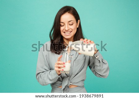 Photo of attractive lady pours milk in glass, dairy products. Wears grey shirt, isolated turquoise color background