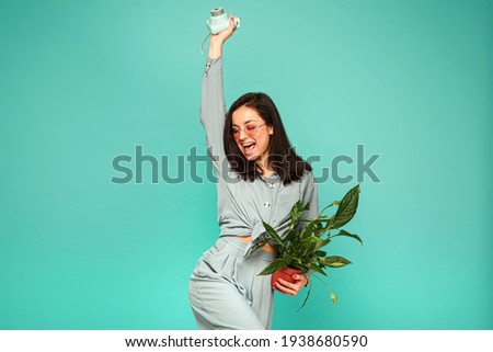 Photo of attractive lady in sunglasses holds camera, plant and has some fun. Wears grey shirt, isolated turquoise color background