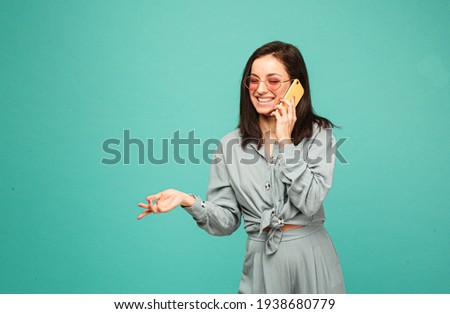 Photo of attractive lady in sunglasses talks on phone and smile. Wears grey shirt, isolated turquoise color background