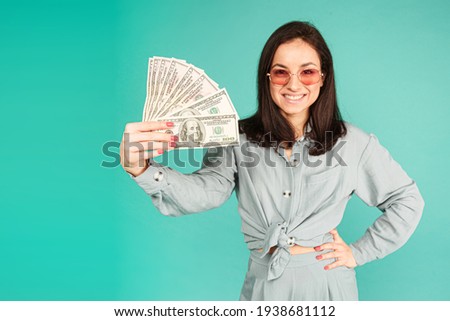 Photo of attractive lady in sunglasses holds money. Wears grey shirt, isolated turquoise color background