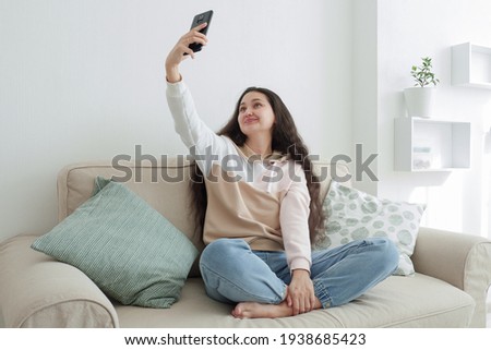Charming Asian girl influencer makes pictures of herself on the phone at home while sitting on the couch. A girl with long curly hair, blue jeans and a beige hoodie shoots content for a blog. Blogging