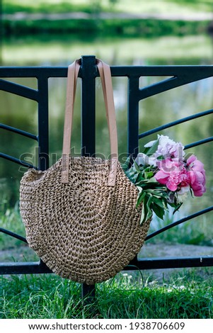 A beige women's wicker bag with a bouquet of pink peonies weighs on the fence of the park against the background of green grass and water near the lake.