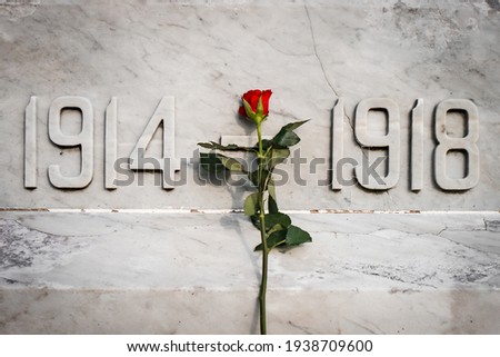 Single red rose flower stood on white marble world war memorial statue 1914 to 1918 first and second remembrance poppy day veterans anniversary. Close up of carved date numbers in stone.