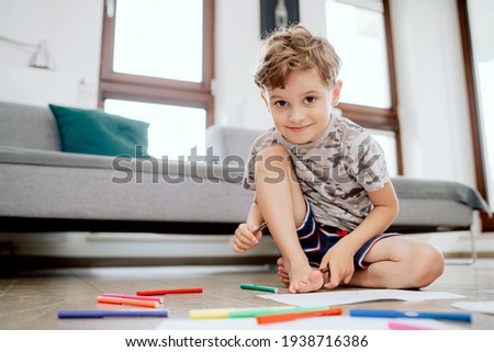 Portrait of a school-aged boy sitting on a wooden floor in the living room of a house. The boy holds a marker in his hand and looks into the camera.
