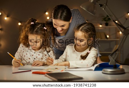 Portrait of mother interested in twins daughters evening activity before bedtime. Affectionate mom looking at preschool children drawing picture and writing. Cozy home atmosphere, quarantine pastime