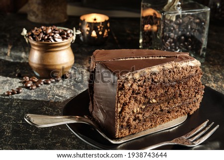 A fancy piece of chocolate cake (Sacher Torte) on a cake slice, a copper jar of coffee beans and a illuminated candel holder are in the out of focus area in the backtrop