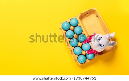 Package of ten blue painted eggs with fluffy white toy bunny on yellow background with copy space, empty text place. Banner for a religion holiday. Design mockup. Fun Easter hunt card. Handmade decor.