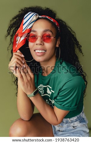 attractive black african american woman in stylish hipster outfit t-shirt and shorts on green studio background, summer fashion trend, happy smiling curly hair red sunglasses accessories