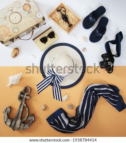 travelling flat lay. female white straw hat on a white and earth tone background. book, sunglasses above, blue towel, photo camera, wooden anchor, box with amphora, vintage suitcase, blue flip flops. 