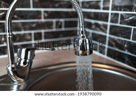 Close-up picture of silver coloured opened faucet, with pouring water in the sink, in the kitchen area. On the background of dark brick wall.