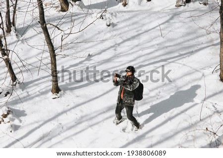 A young male tourist in black warm clothes and a large backpack on his back walks through the snowy forest and takes pictures of nature on a mobile phone.