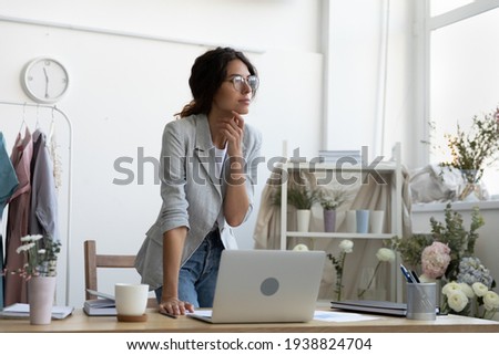 Dreamy young Caucasian female florist look in distance distracted from computer work thinking or planning. Happy businesswoman use laptop make decision, visualize or dream in cozy home office. Royalty-Free Stock Photo #1938824704