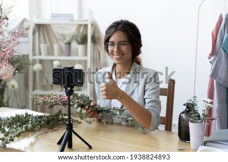 Smiling young Caucasian female designer show thumb up shoot vlog recommend floral design course or training. Happy woman florist record video blog or tutorial on smartphone of flower decor making. Royalty-Free Stock Photo #1938824893