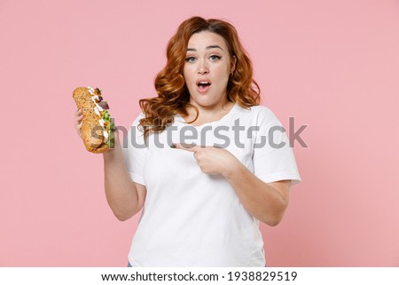 Shocked young redhead plus size body positive woman 20s in white casual t-shirt pointing index finger on american classic fast food hot dog isolated on pastel pink color background studio portrait