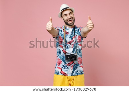 Funny young traveler tourist man in summer basic clothes hat with photo camera showing thumbs up like gesture isolated on pink background. Passenger traveling on weekends. Air flight journey concept