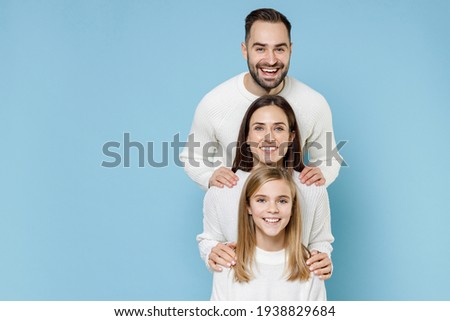 Smiling young happy parents mom dad with child kid daughter teen girl in basic white sweaters stand behind each other isolated on blue color background studio portrait. Family day parenthood concept