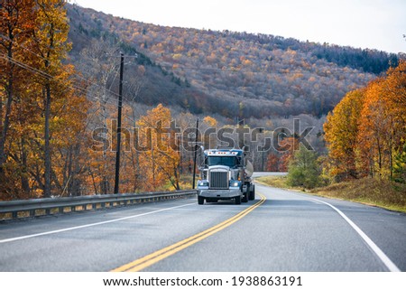 Classic blue day cab industrial grade professional big rig semi truck transporting fuel in tank semi trailer climbing uphill on the winding road in autumn mountain in Vermont New England Royalty-Free Stock Photo #1938863191