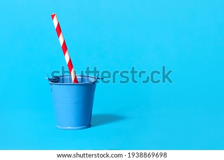 One blue toy bucket on a light blue background with a striped red and white drinking straw. Weekend party concept with non-standard serving drinks. Free space for text