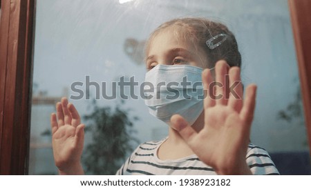 sad kid girl in protective medical mask by the window looks out. stay home coronavirus covid 19 concept. sad child in protective mask in hospital pandemic looking out window. stay home coronavirus
