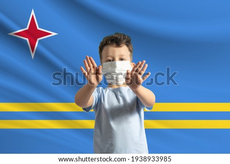 Little white boy in a protective mask on the background of the flag of Aruba. Makes a stop sign with his hands, stay at home Aruba.
