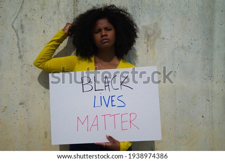 angry african-american woman with raised fist holds up a banner reading black lives matter. In the background grey wall.