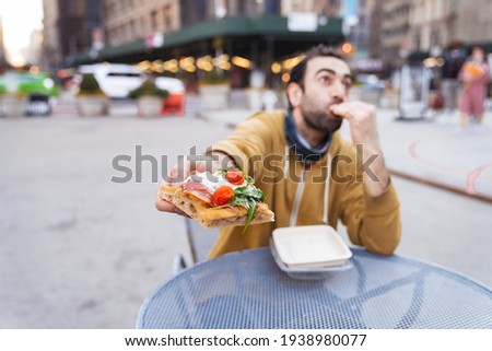 Young man having pizza snack outdoors, sitting on the table, showing emotions. MIddle eastern adult. Wide angle shot