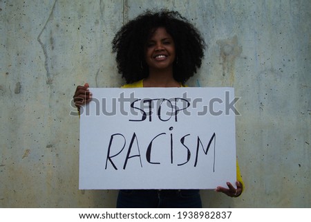 african-american woman holds up a banner reading STOP RACISM. In the background grey wall.
