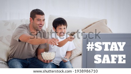 Stay safe text over father and son watching television and eating popcorn. covid 19 pandemic and social distancing concept digitally generated image.