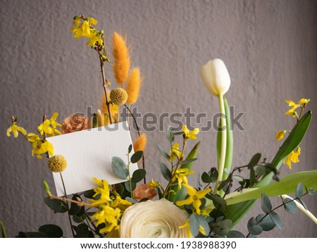 Blank greeting card on a bouquet of spring flowers, white ranunculus, sprigs of yellow forsythia and eucalyptus, pink and white tulips, orange dianthus in a glass vase on a gray concrete background in