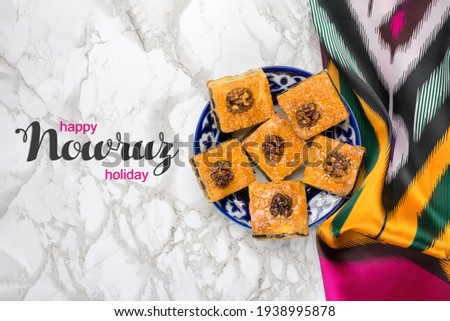 Traditional arabic dessert baklava with walnuts, raisins on plate, with Uzbek national ornament, Ikat fabric on white wooden table Text Happy Nowruz Holiday Concept of spring came Top view Flat lay